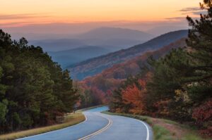 A gently curving road leading into a forested mountain range with a sunset in the background
